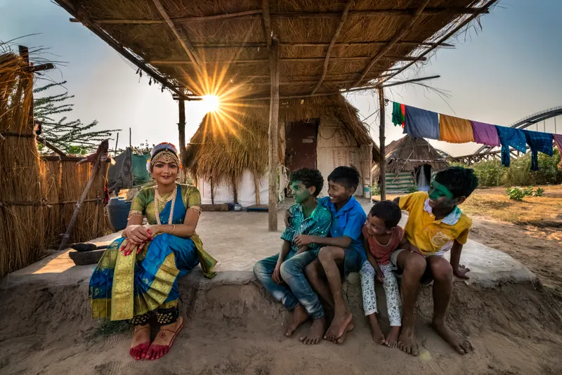 A dancer in full costume resting with village kids under a thatched shade