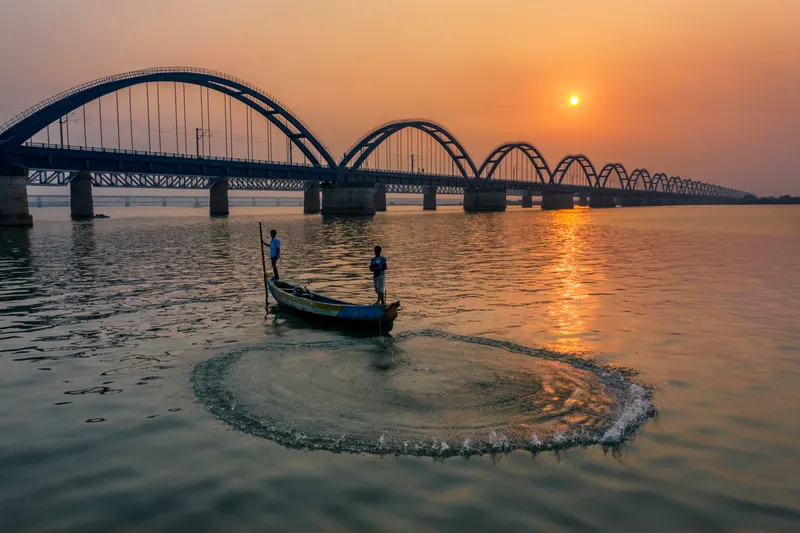 Fishermen casting a net from a boat below the arch bridge at sunset