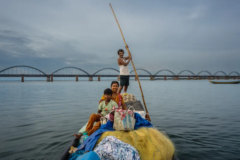 A fisherman poling his family’s boat, piled with nets, across the Godavari with the arched rail bridge behind