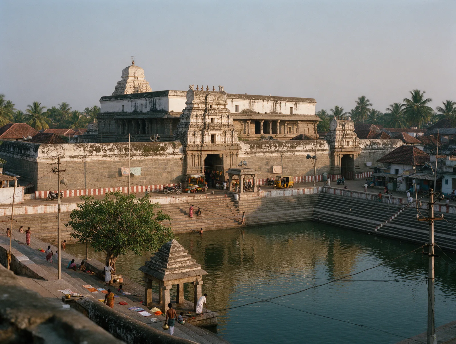 The Bhimeswara temple at Draksharamam seen across the Sapta Godavari tank at golden hour, devotees on the stepped ghat, the weathered granite wall and five-tiered gopuram beyond, and the white upper storey of the two-storeyed shrine rising behind.