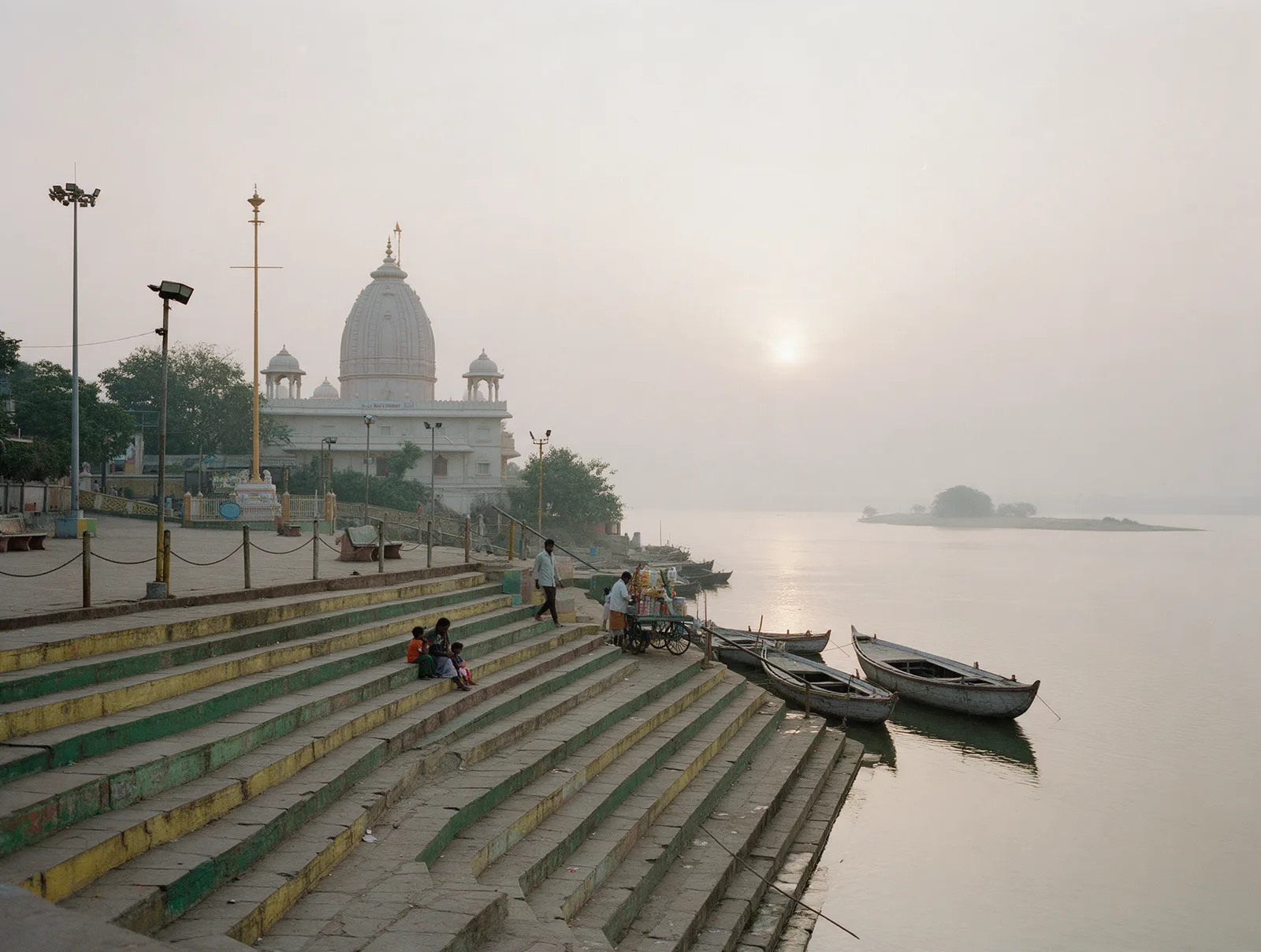 Warm late-afternoon haze over the wide Godavari from the weathered stone steps of Gowthami Ghat in Rajamahendravaram, with the pale domed ISKCON temple among trees along the bank.