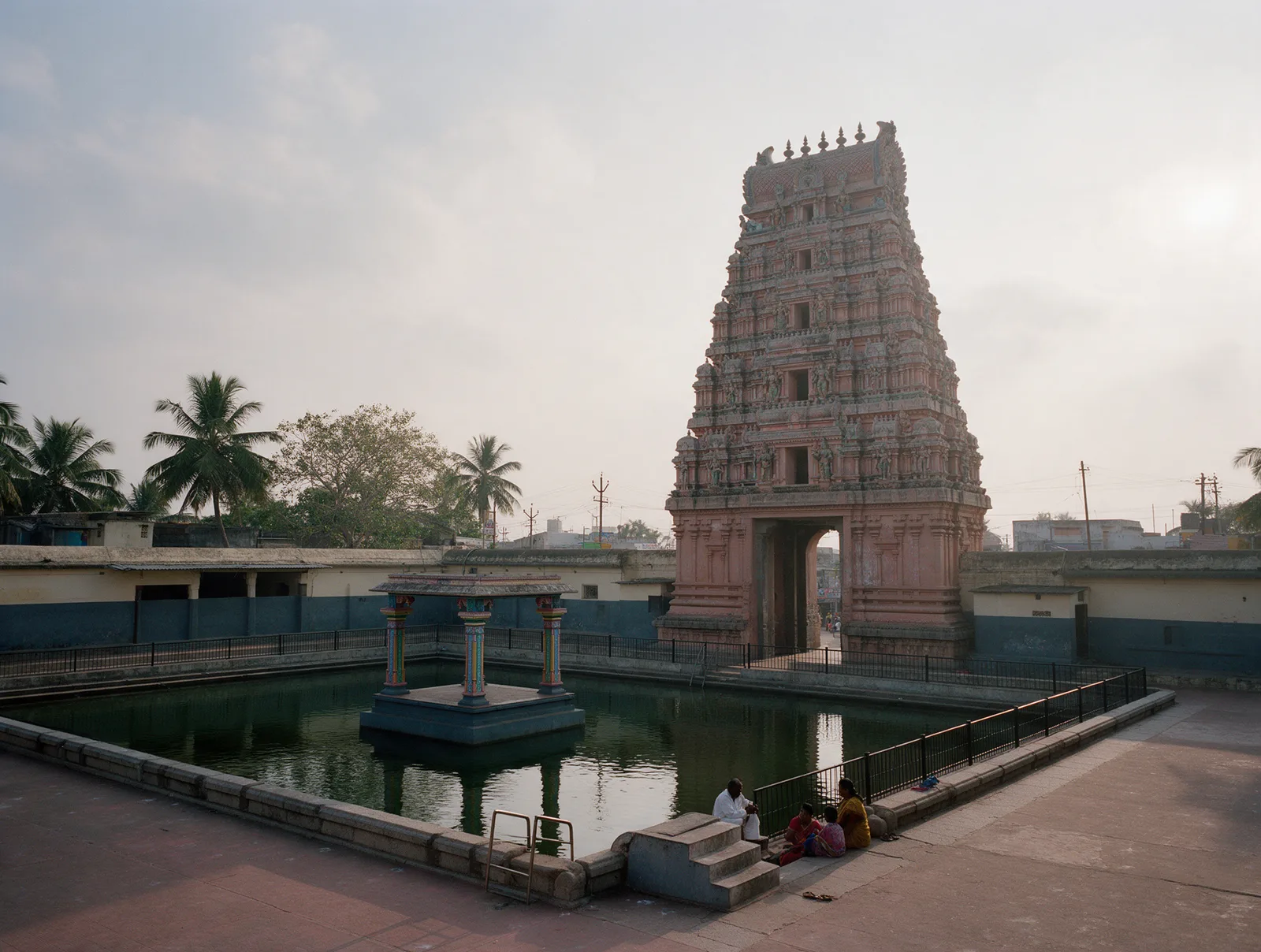 The tall, slim five-tier north gopuram of the Kukkuteswara temple at Pithapuram, an arched passage through its base, rising at the head of the dark green Pada Gaya tank, with a small polychrome-pillared pavilion in the water under a hazy delta sky