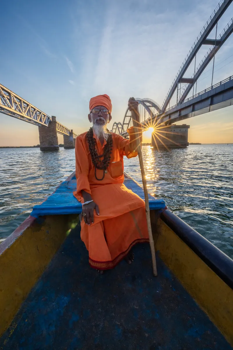 A sadhu in saffron seated at the bow of a boat between the Godavari bridges at sunset